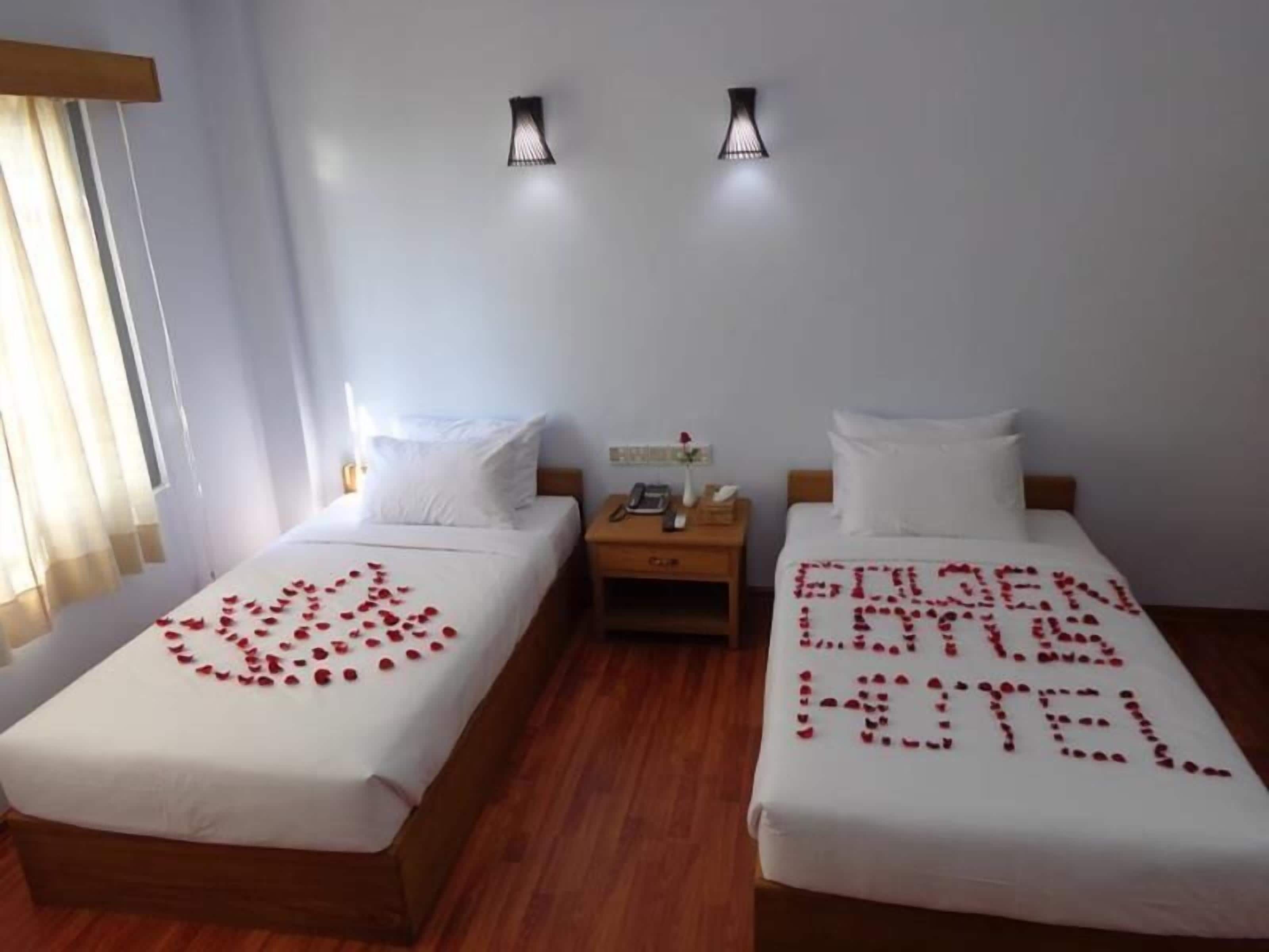 A hotel room with two beds, a nightstand with a phone and a vase with a flower, and a wall-mounted light fixture.