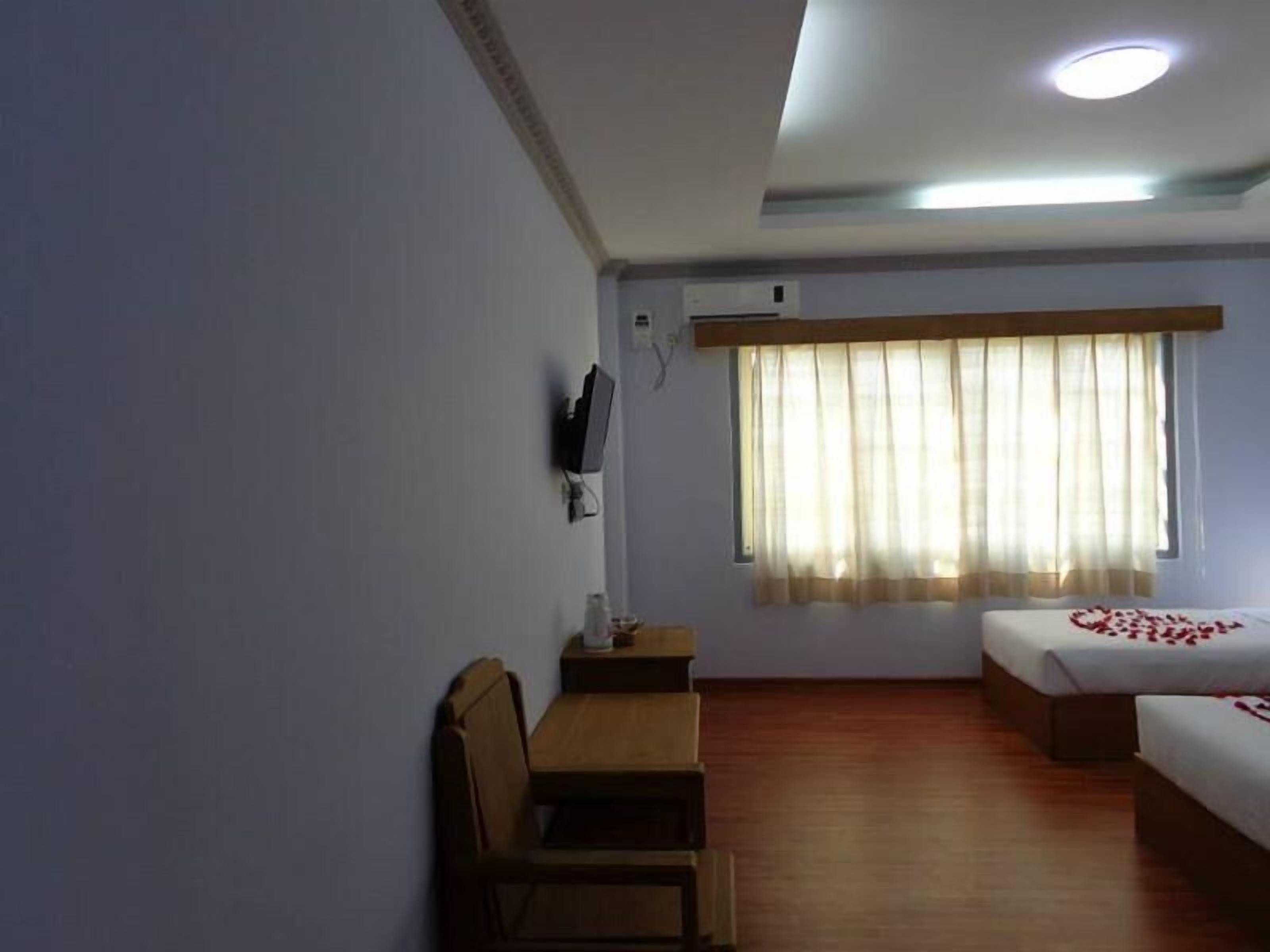 Une chambre d’hôtel avec deux lits simples, un bureau en bois, une chaise, un téléviseur fixé au mur et une fenêtre avec des rideaux légers.