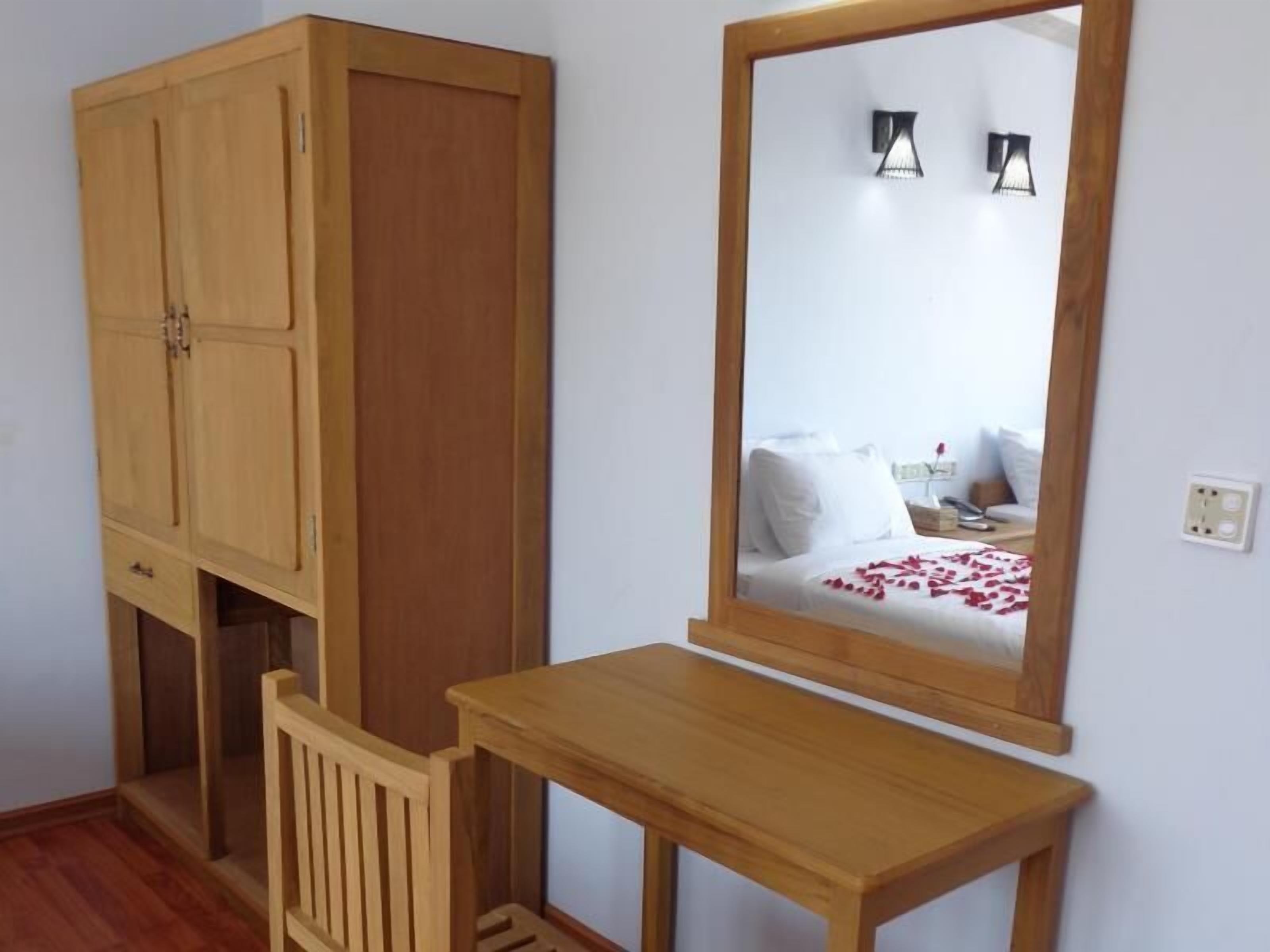 A bedroom with a wooden wardrobe, a bedside table, and a mirror reflecting a bed with white bedding and a red rose.