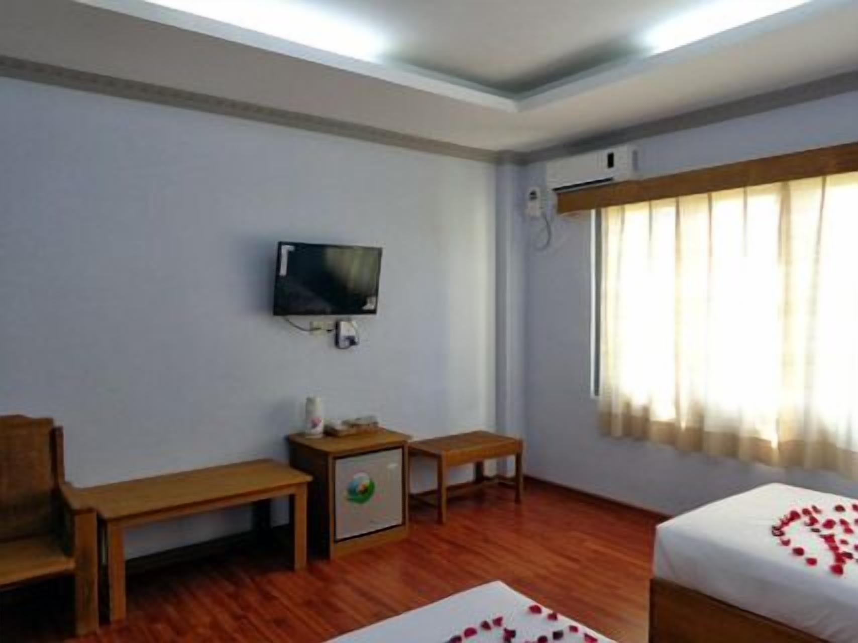 A hotel room with a television, a small cabinet, a wooden desk, a wooden chair, and a bed with a heart-shaped decoration on the bedspread.