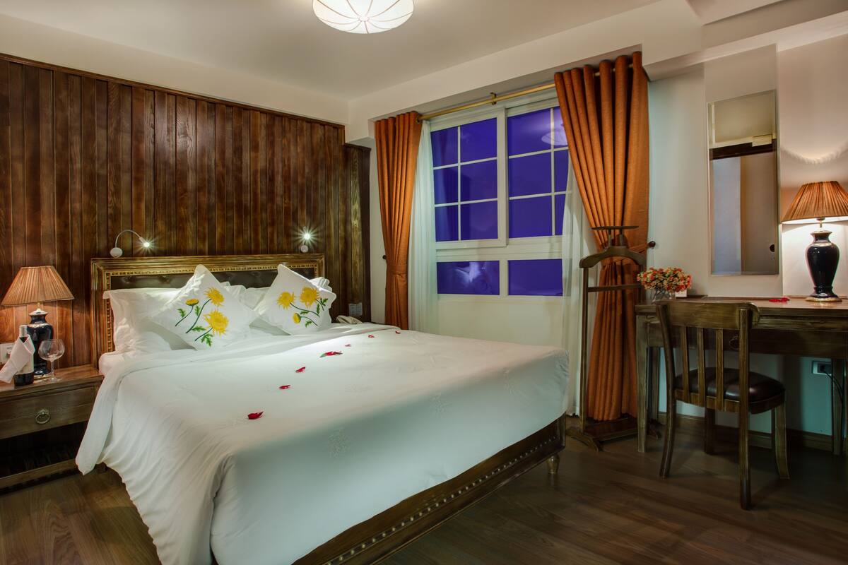 Hotelkamer met een groot bed, houten lambrisering, een bureau met stoel en een raam met gordijnen.