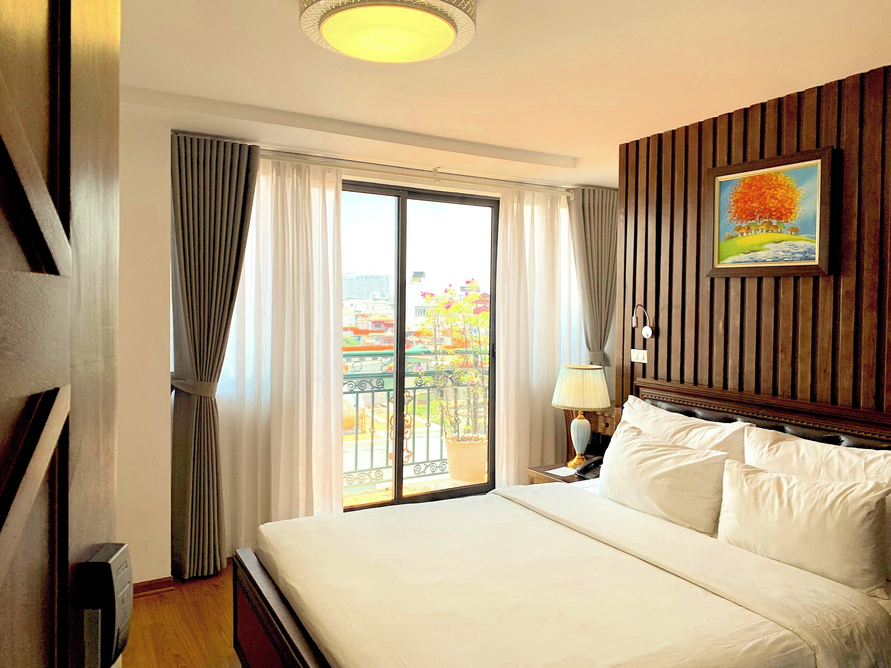 A hotel room with a large bed, a nightstand, a lamp, a painting, and a view of a balcony with a railing.