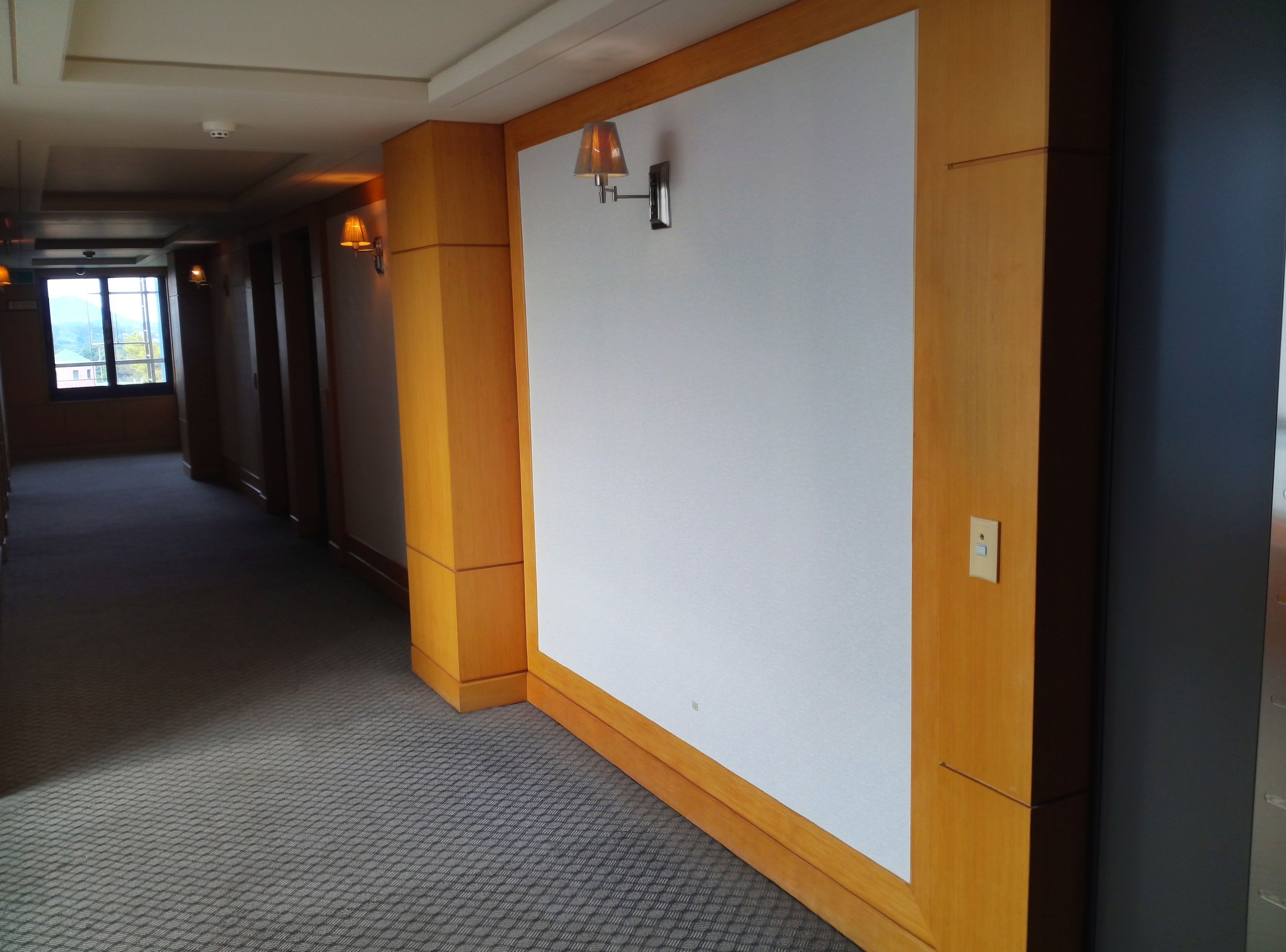 A hotel corridor with wooden paneling, a large white wall, and a window at the end.