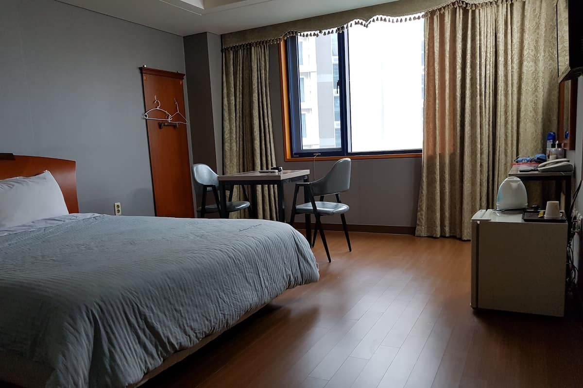 A hotel room with a bed, a desk, two chairs, a small table with a kettle, and a window with curtains.