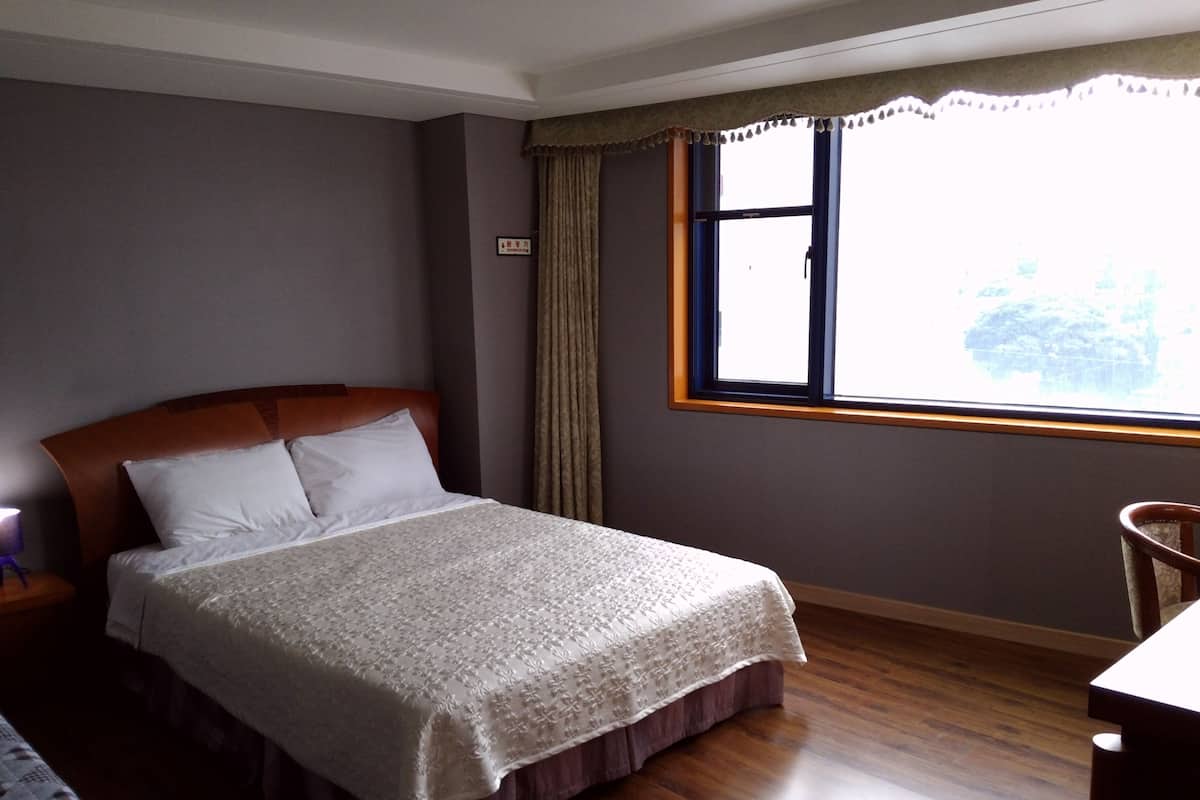 A neatly made bed with a wooden headboard, a window with curtains, and a wooden floor.
