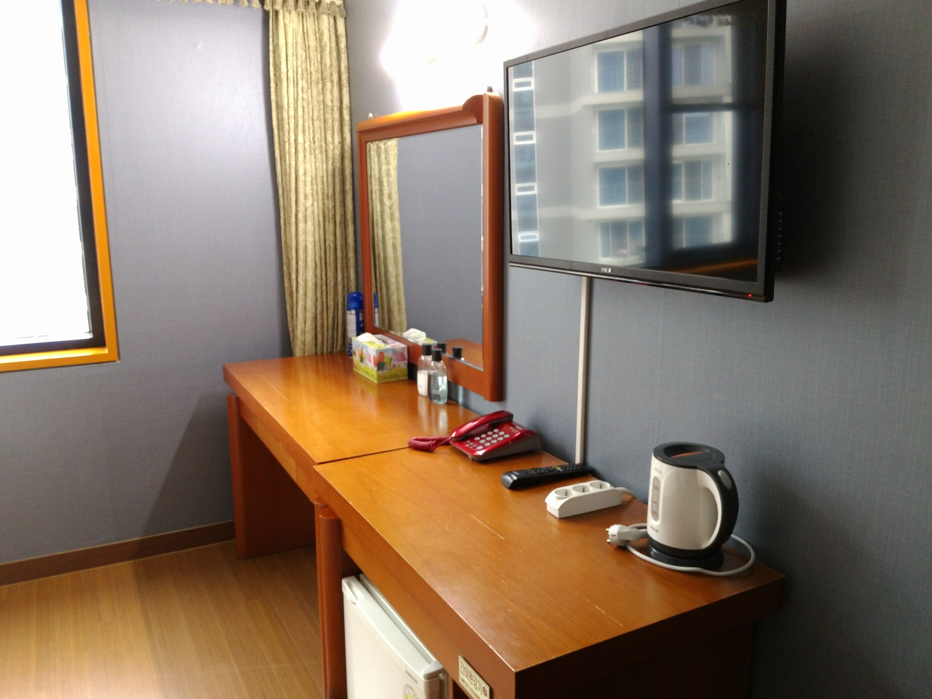 A hotel room with a wooden desk, a flat-screen TV mounted on the wall, a mirror, a telephone, a remote control, a kettle, and a small refrigerator.