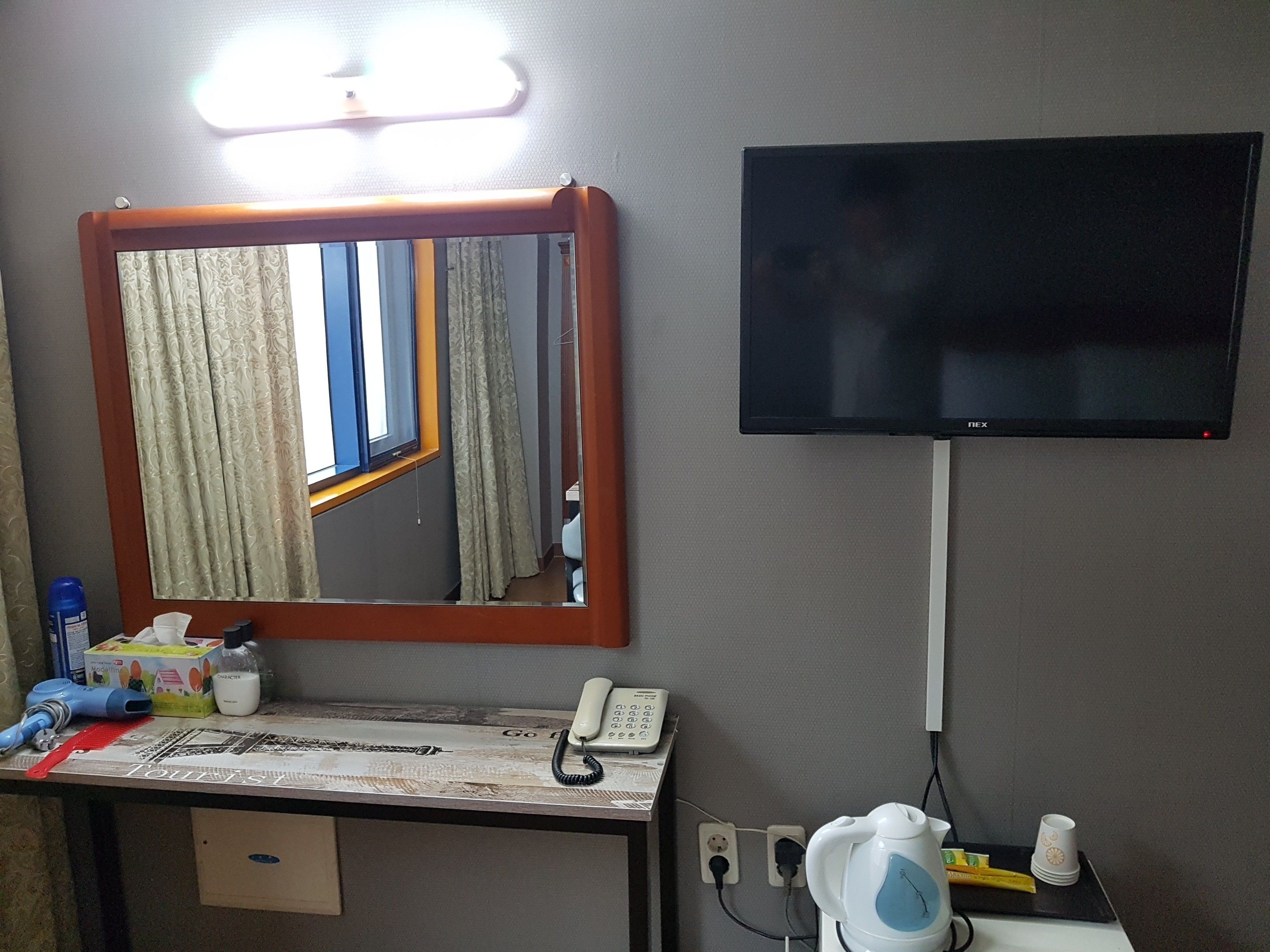 A hotel room with a large mirror, a flat-screen TV mounted on the wall, a desk with a telephone, a kettle, and a cup.