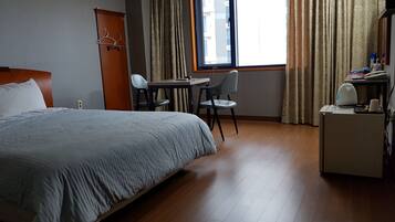 A hotel room with a bed, a desk, two chairs, a small table, a kettle, and a window with curtains.