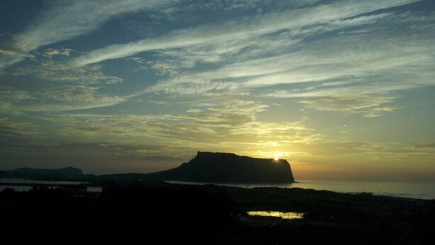 Un coucher de soleil sur un paysage côtier, avec une falaise imposante et un petit plan d'eau au premier plan.
