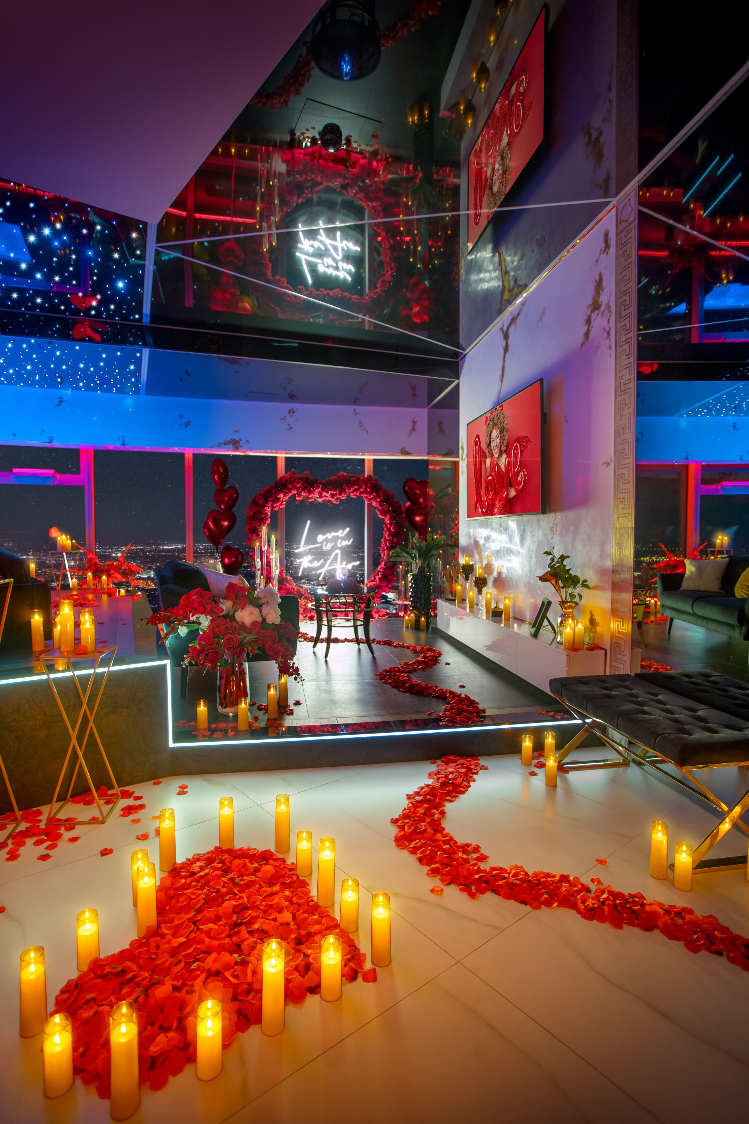 A romantic indoor setting with a heart-shaped aisle of rose petals and candles, a seating area with a red heart arch, and a city view through large windows.