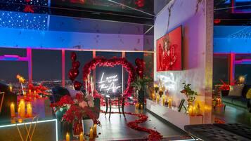 A romantic indoor setting with a heart-shaped aisle of rose petals and candles, a seating area with a red heart arch, and a city view through large windows.