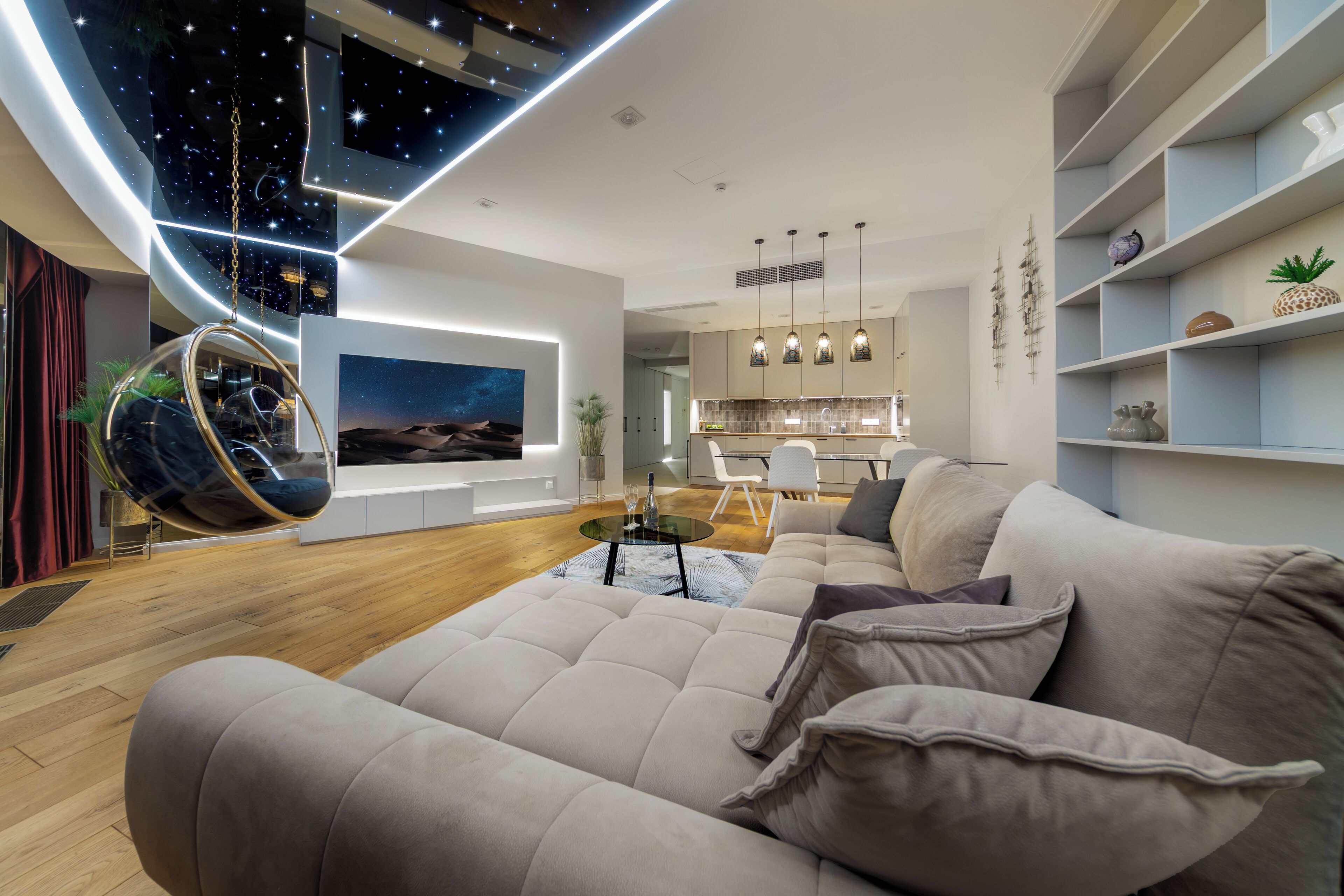 Un salon moderne avec un grand canapé, un fauteuil suspendu, un téléviseur à écran plat et un coin cuisine à l’arrière-plan.