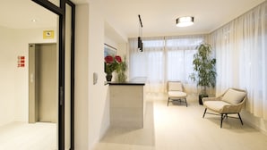 Un salon moderne avec un plancher en bois, un îlot central agrémenté d’un vase de fleurs, deux fauteuils en osier, une grande plante en pot et une porte-fenêtre coulissante donnant sur une autre pièce.