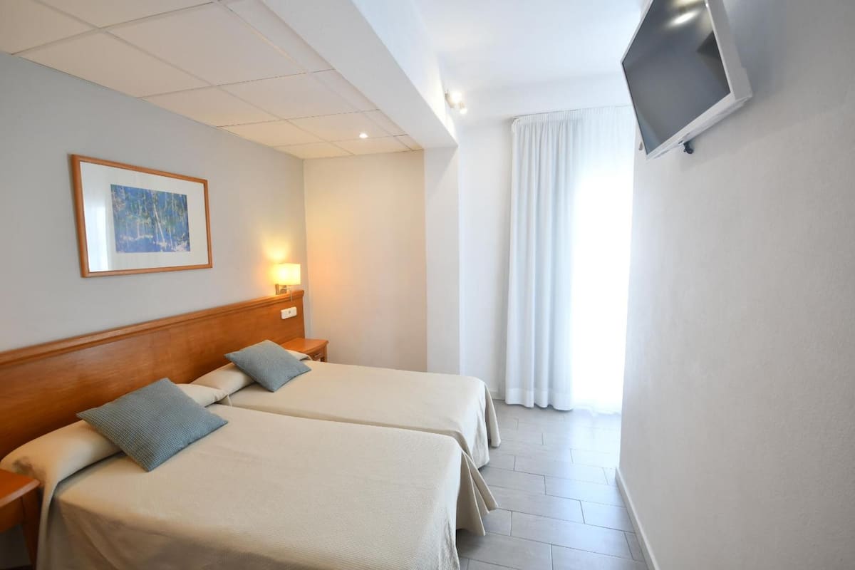 Ein Hotelzimmer mit zwei Betten, einem an der Wand befestigten Fernseher, einem gerahmten Bild und einem Fenster mit Vorhängen.