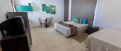 Quarto de hotel com uma cama, uma escrivaninha com televisão, uma cadeira, uma mesinha e uma janela.