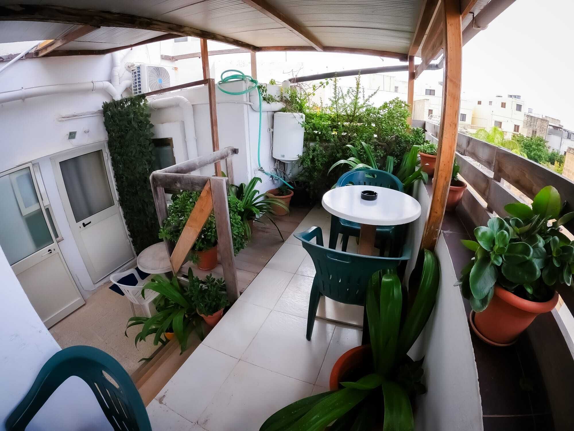 A small balcony with a table and chairs, surrounded by potted plants and a wooden structure.
