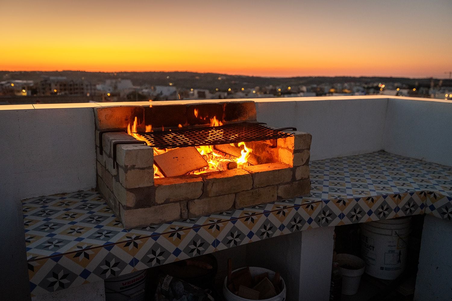 Una parrilla de ladrillo con rejilla de metal, ubicada en un patio con baldosas y vistas a un paisaje urbano.