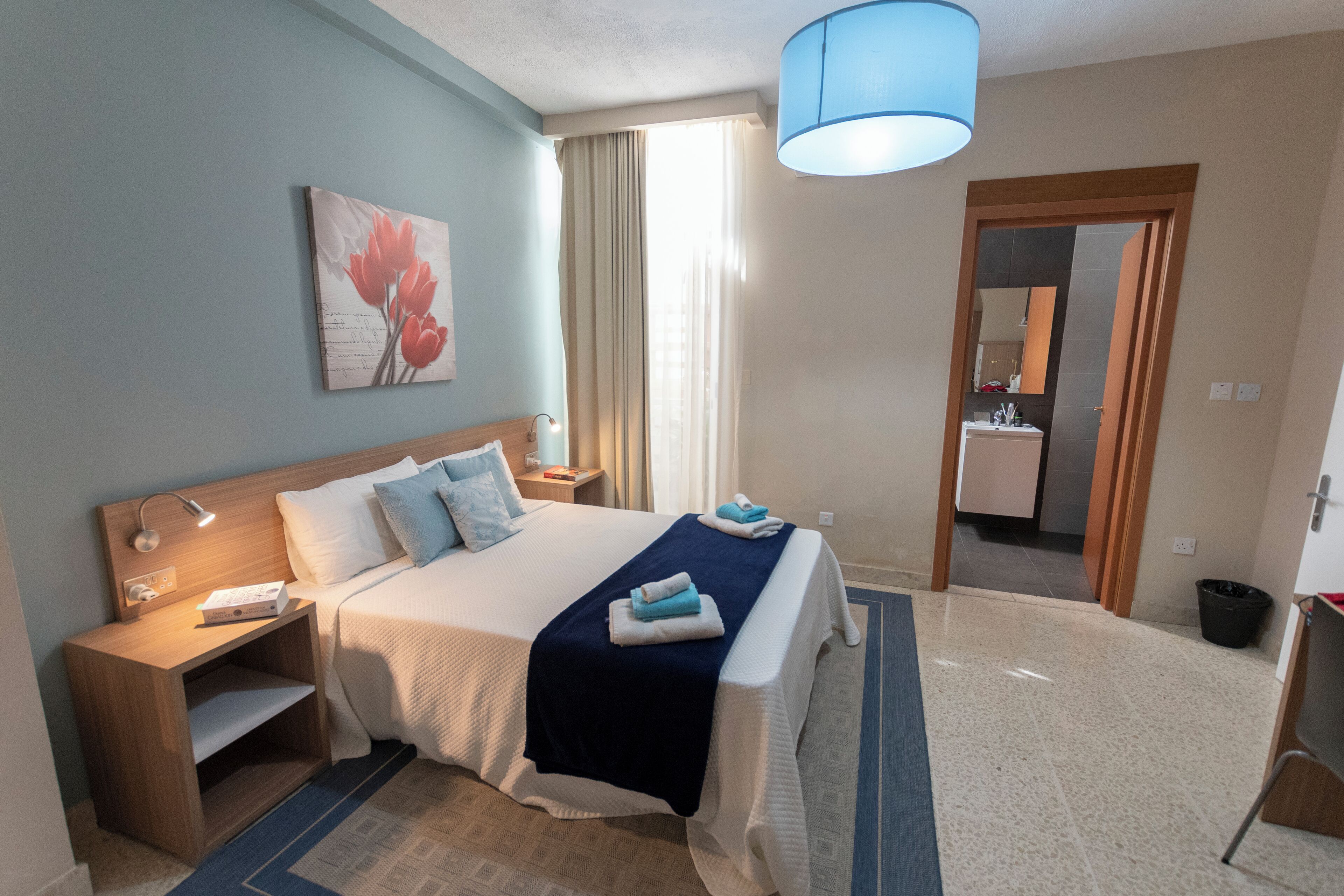 A hotel room with a bed, a nightstand, a lamp, a painting, a bathroom mirror, and a door leading to another room.