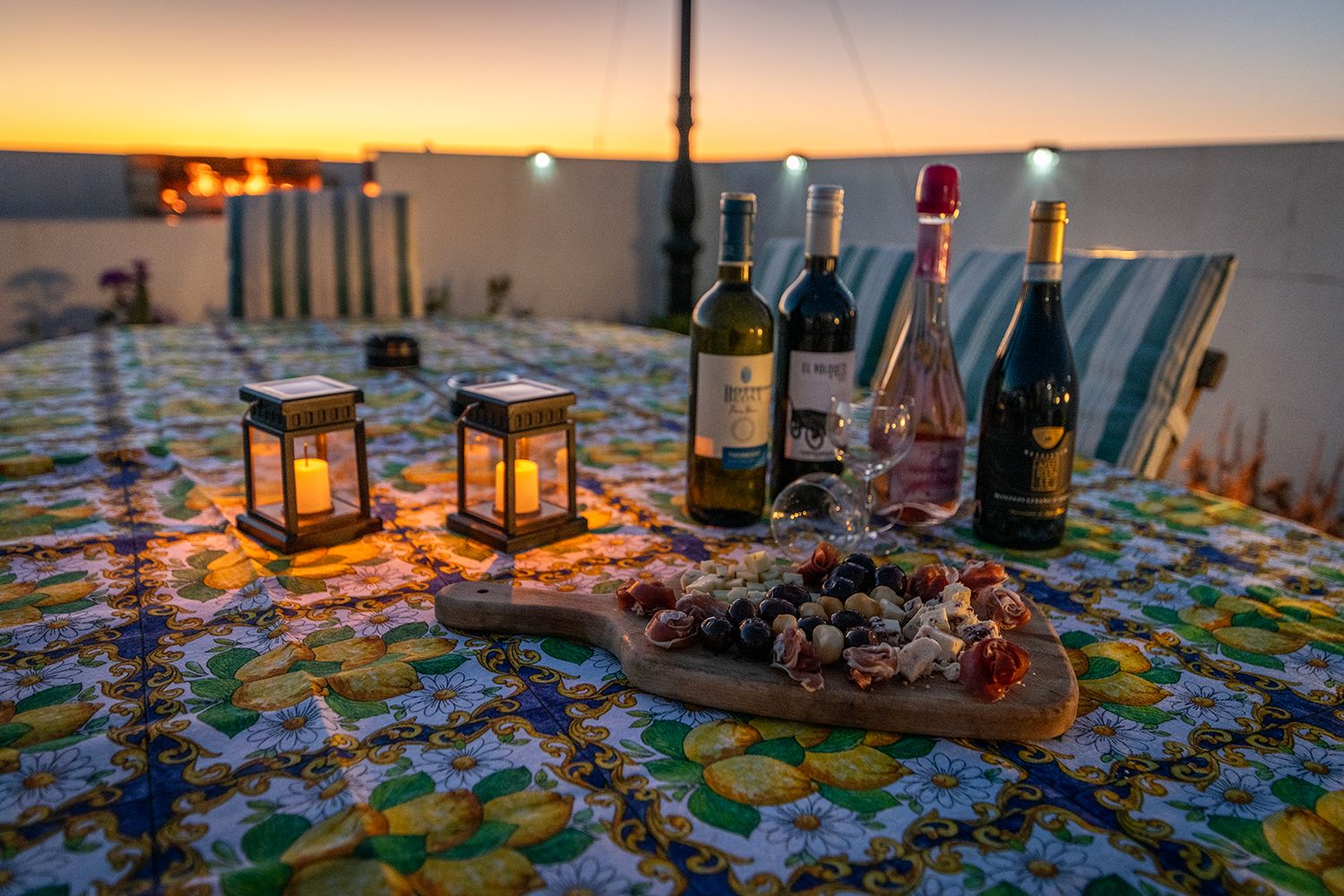 Une table dressée avec des bouteilles de vin, des verres et une planche de fromages, le tout éclairé par des lanternes au coucher du soleil.