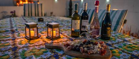Une table dressée avec des bouteilles de vin, des verres et une planche de fromages, le tout éclairé par des lanternes au coucher du soleil.