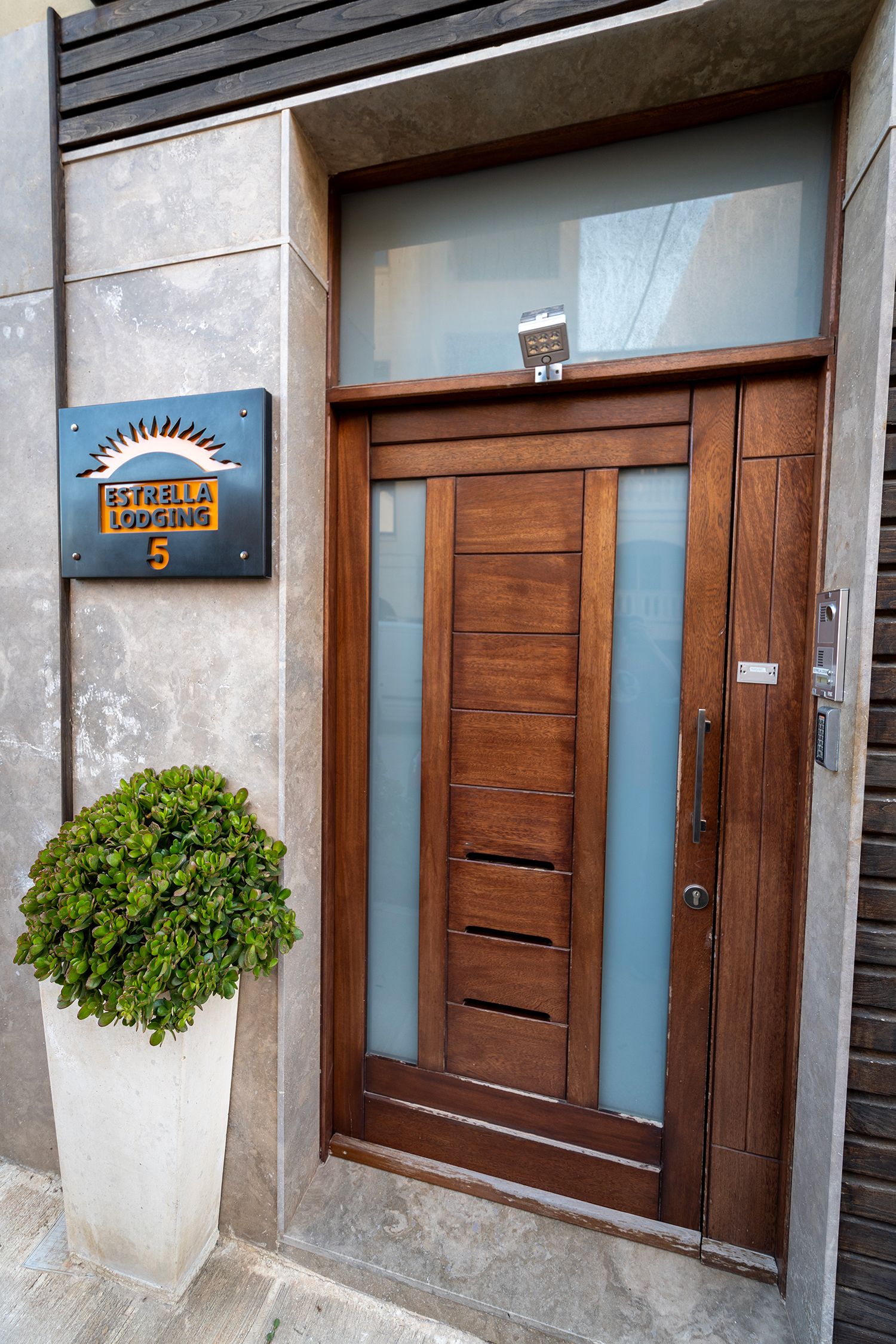 Uma entrada moderna com porta de madeira, uma placa de metal acima e um vaso de planta ao lado da porta.