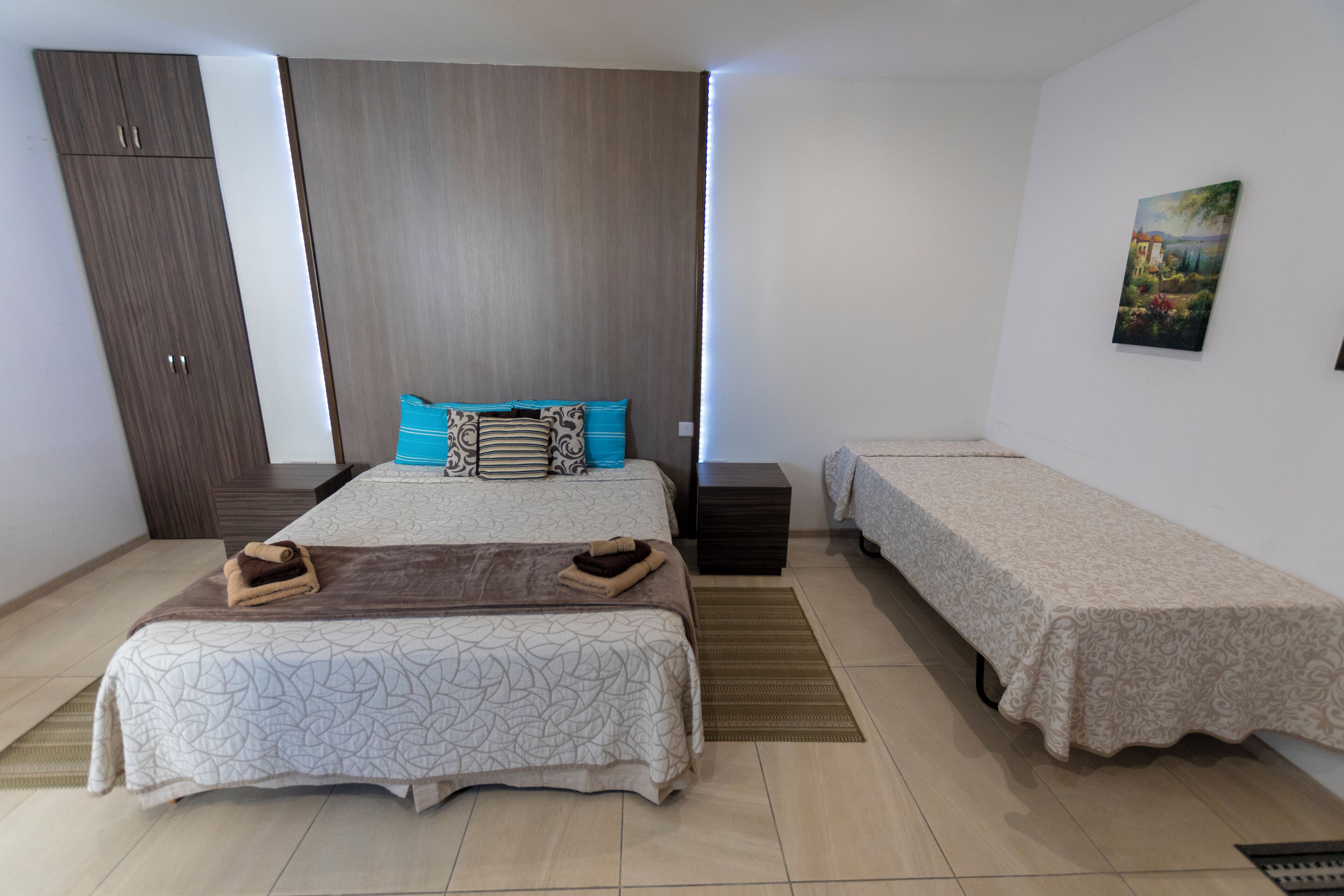 A bedroom with a large bed, a smaller bed, a wardrobe, and a painting on the wall.