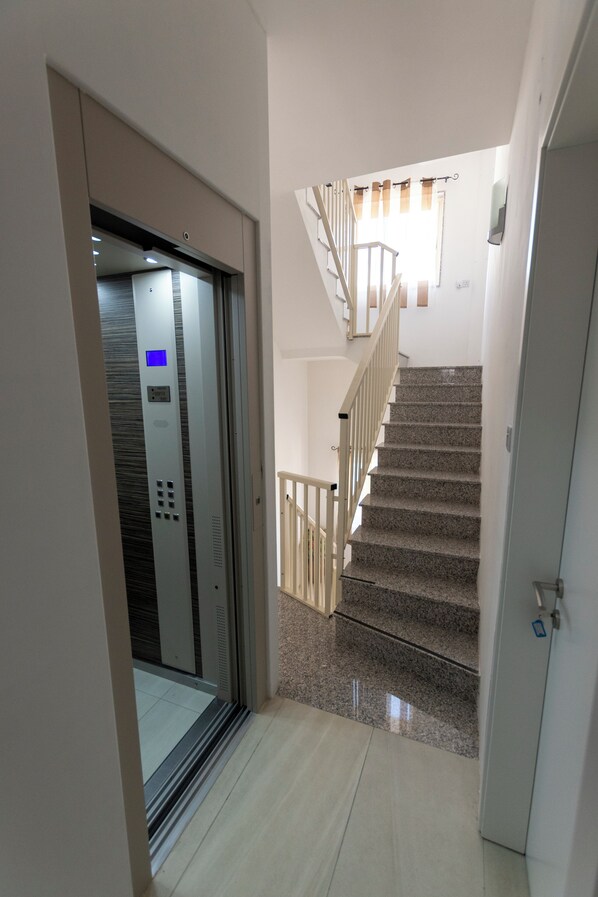 Interior moderno com elevador de vidro, escada com corrimão de madeira e porta com puxador.