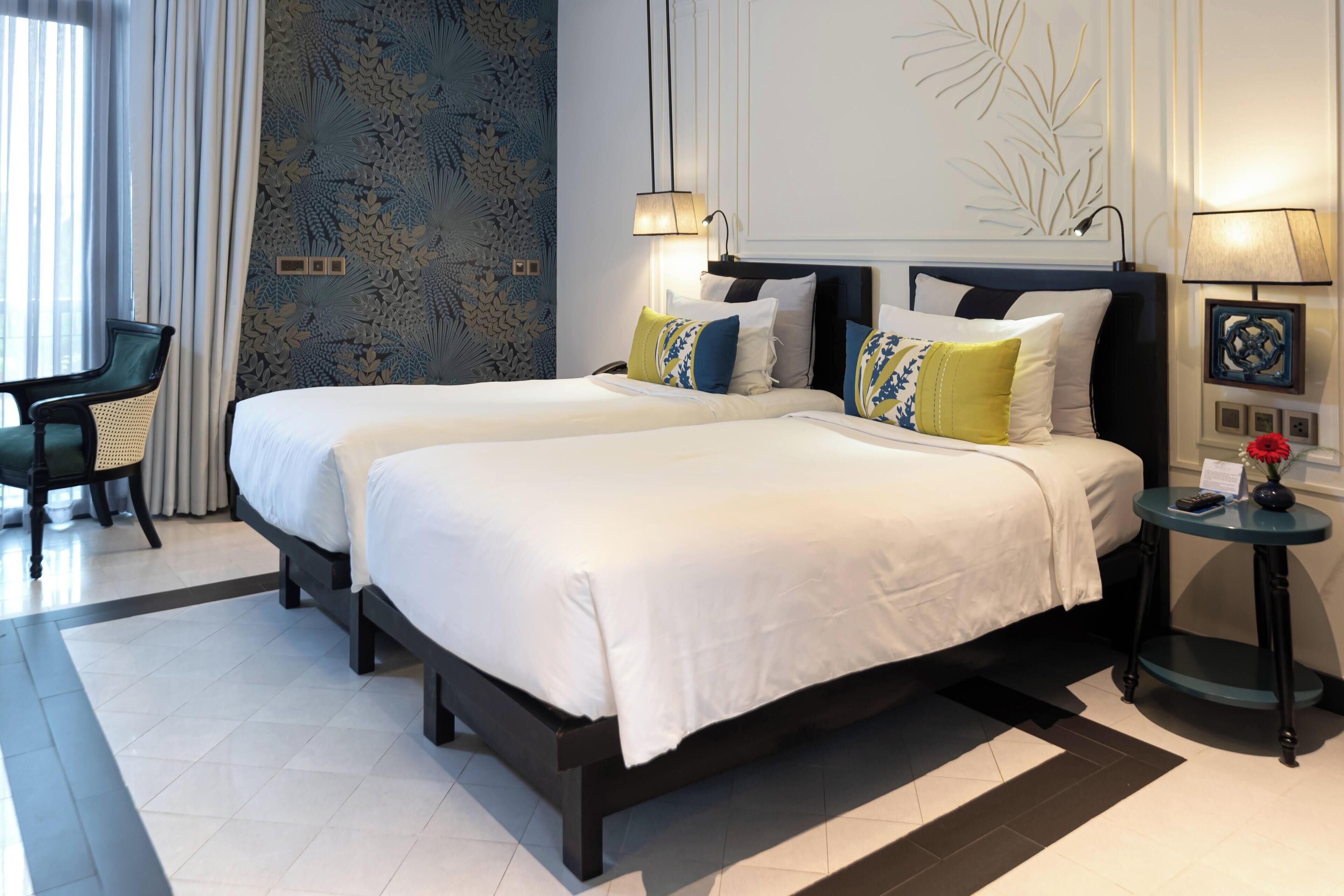 A hotel room with two beds, a small table with a lamp, and a chair.