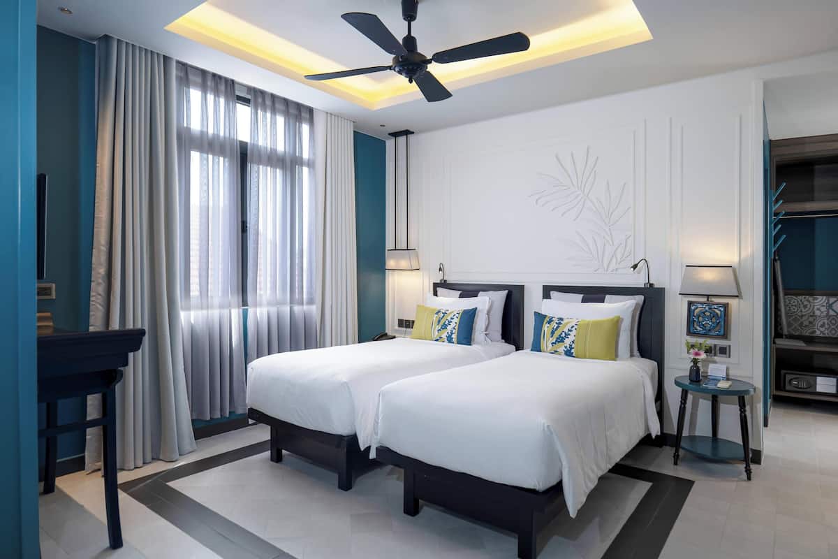 A hotel room with two beds, a ceiling fan, a desk, and a small table with a lamp.