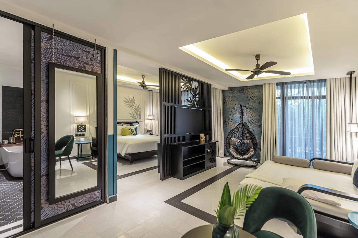 A modern hotel room with a large bed, a sofa, a TV, a ceiling fan, and a large window with curtains.