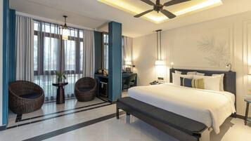 A hotel room with a large bed, two wicker chairs, a small table with a plant, a desk with a lamp, and a ceiling fan.