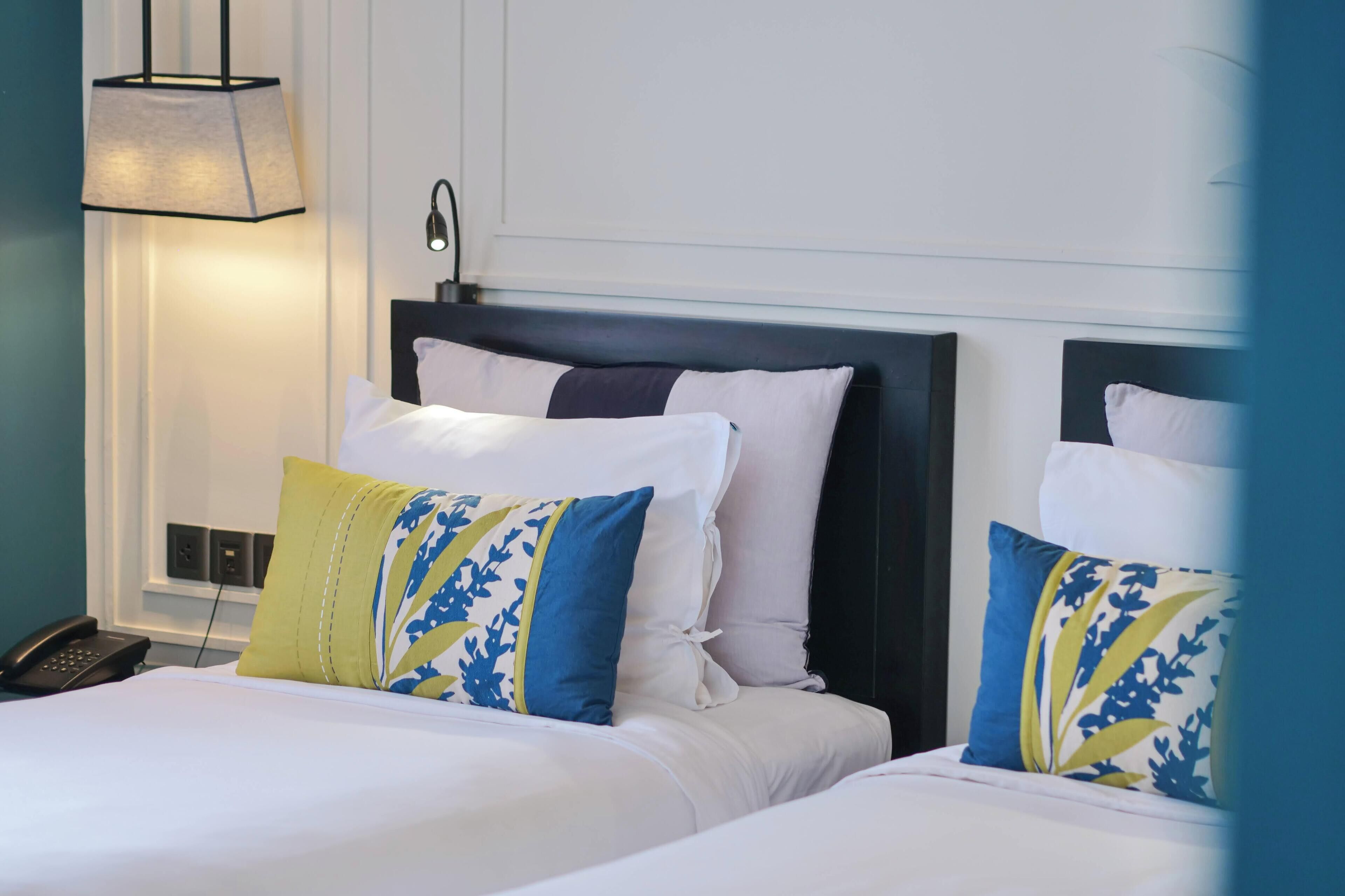 Habitación de hotel con dos camas, cada una con ropa de cama blanca y almohadas estampadas en azul y amarillo. Hay una mesita de noche con una lámpara y un teléfono.