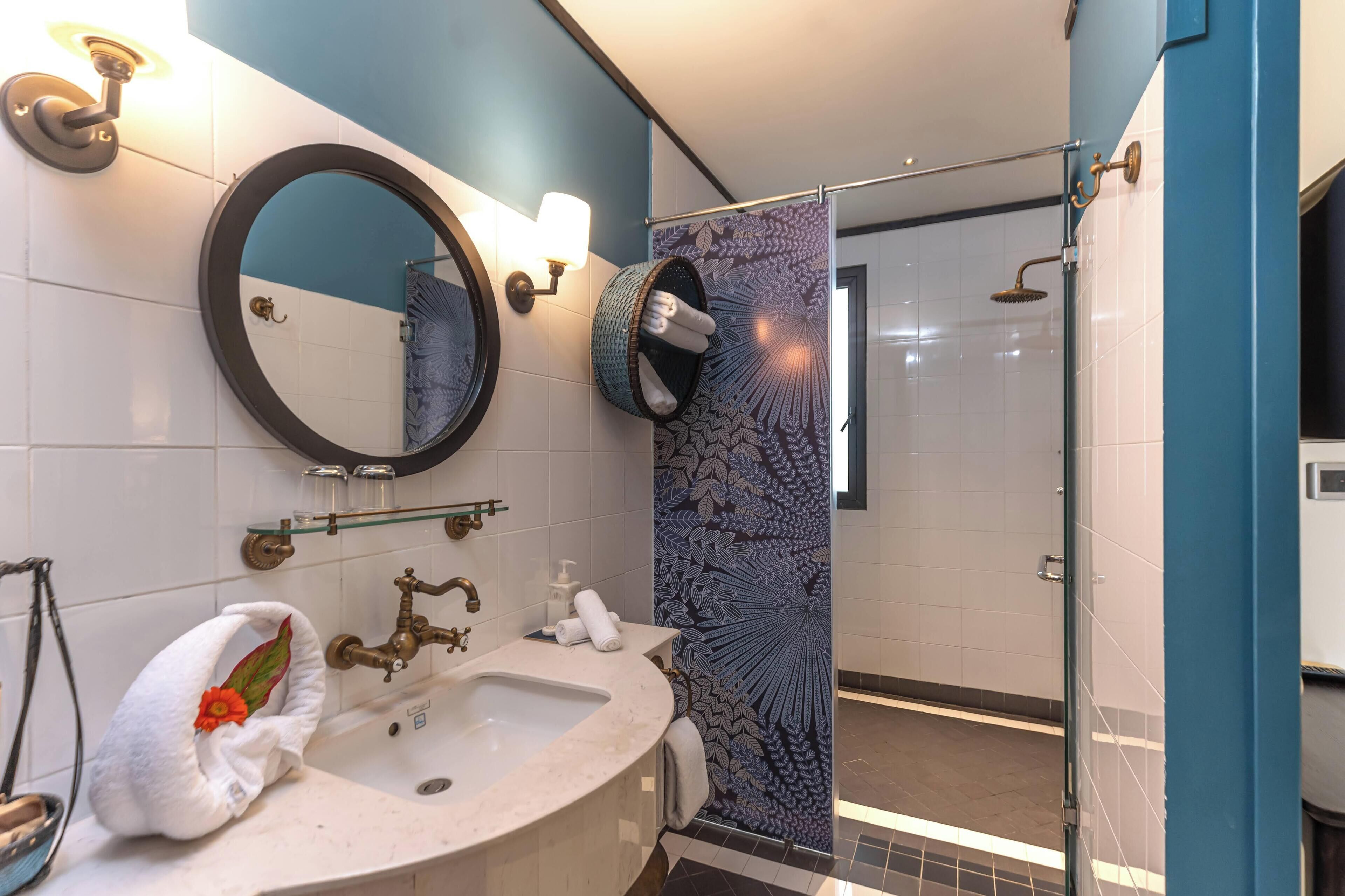 A bathroom with a round mirror, a sink with a towel, a shower with a glass door, and a patterned shower curtain.