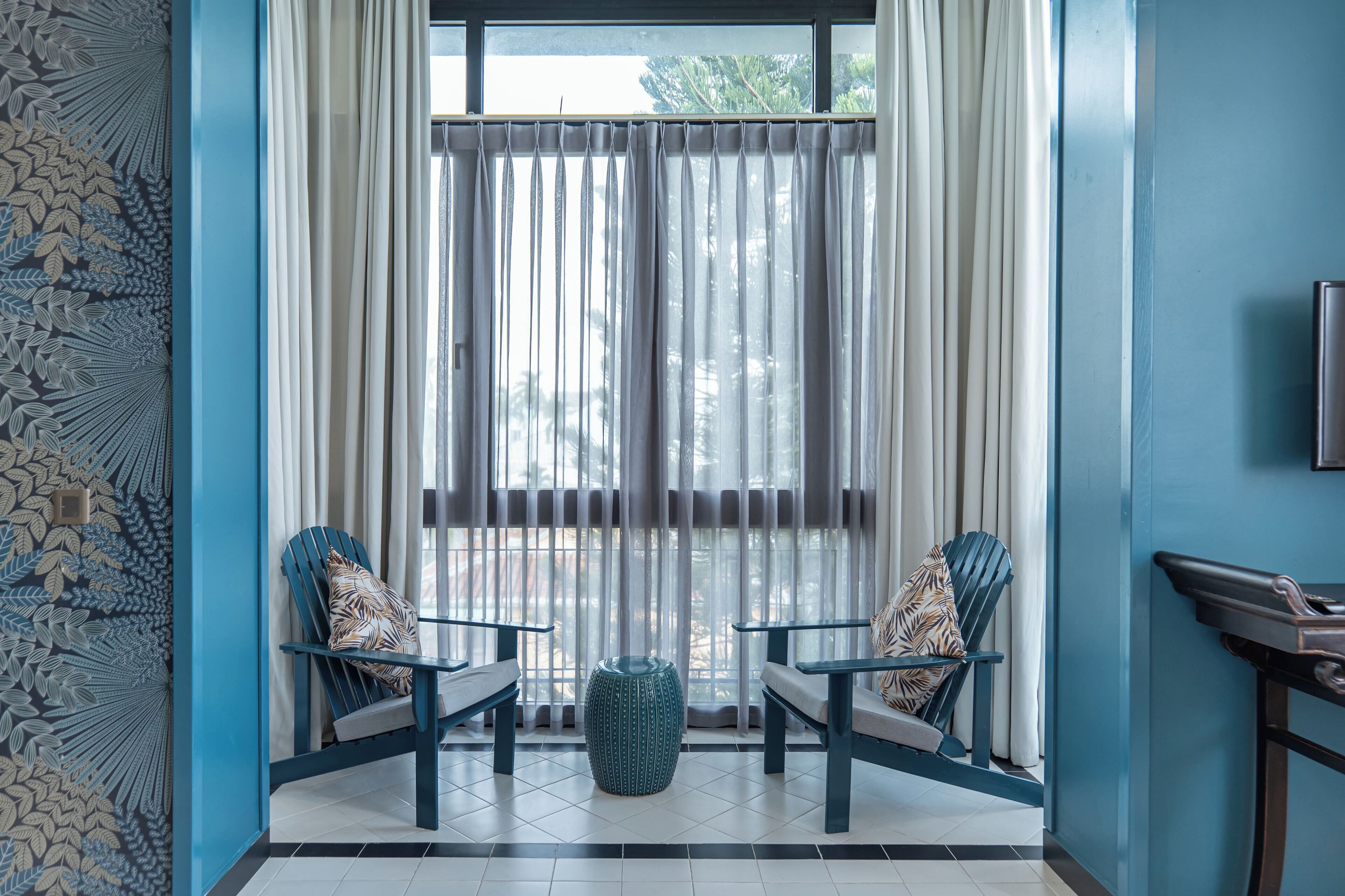 Une pièce avec deux chaises bleues, un petit pouf bleu et une grande fenêtre ornée de rideaux légers.
