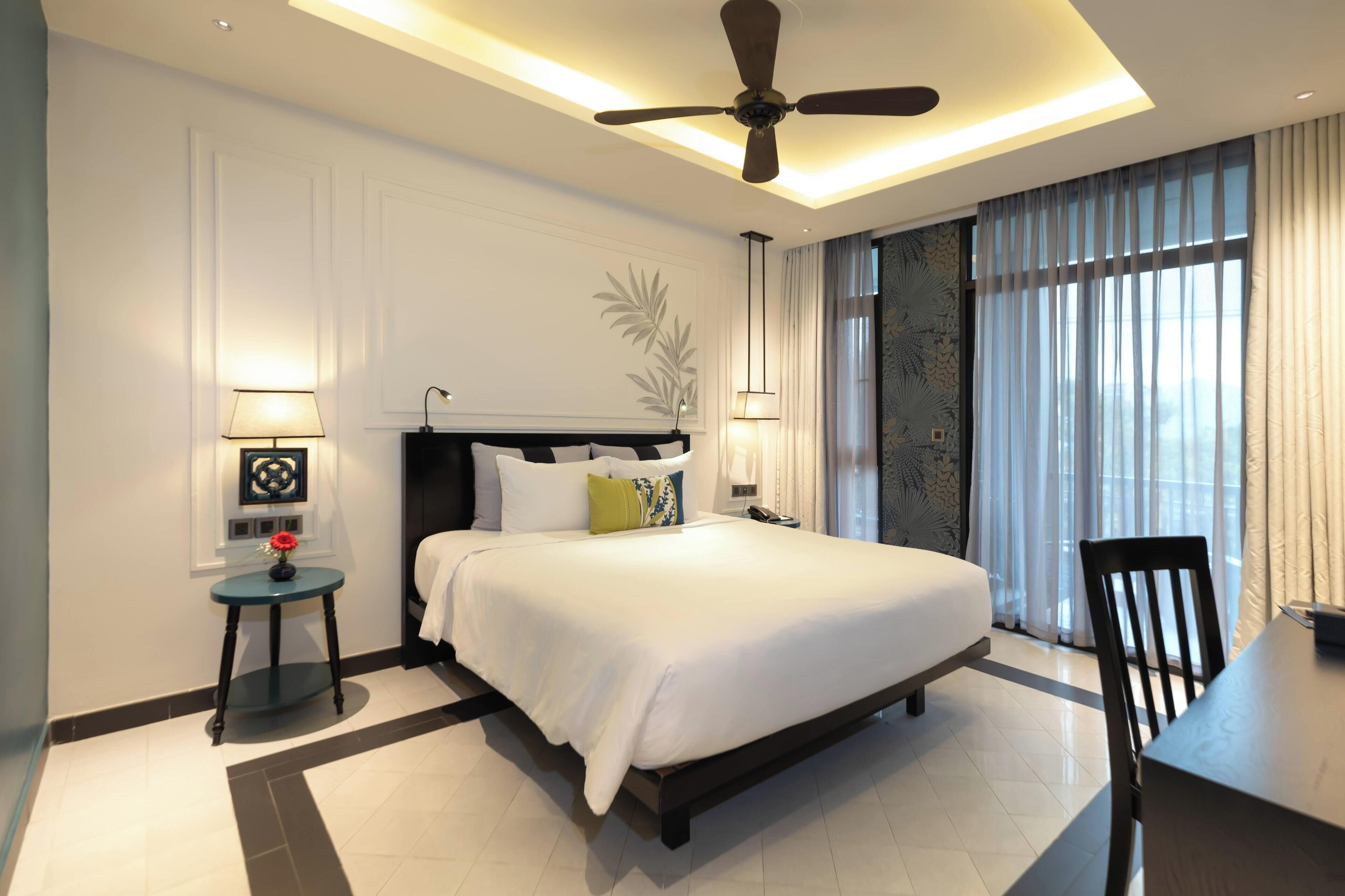 Habitación de hotel moderna con una cama grande, lámparas de noche, ventilador de techo y vistas de la ciudad a través de amplios ventanales.