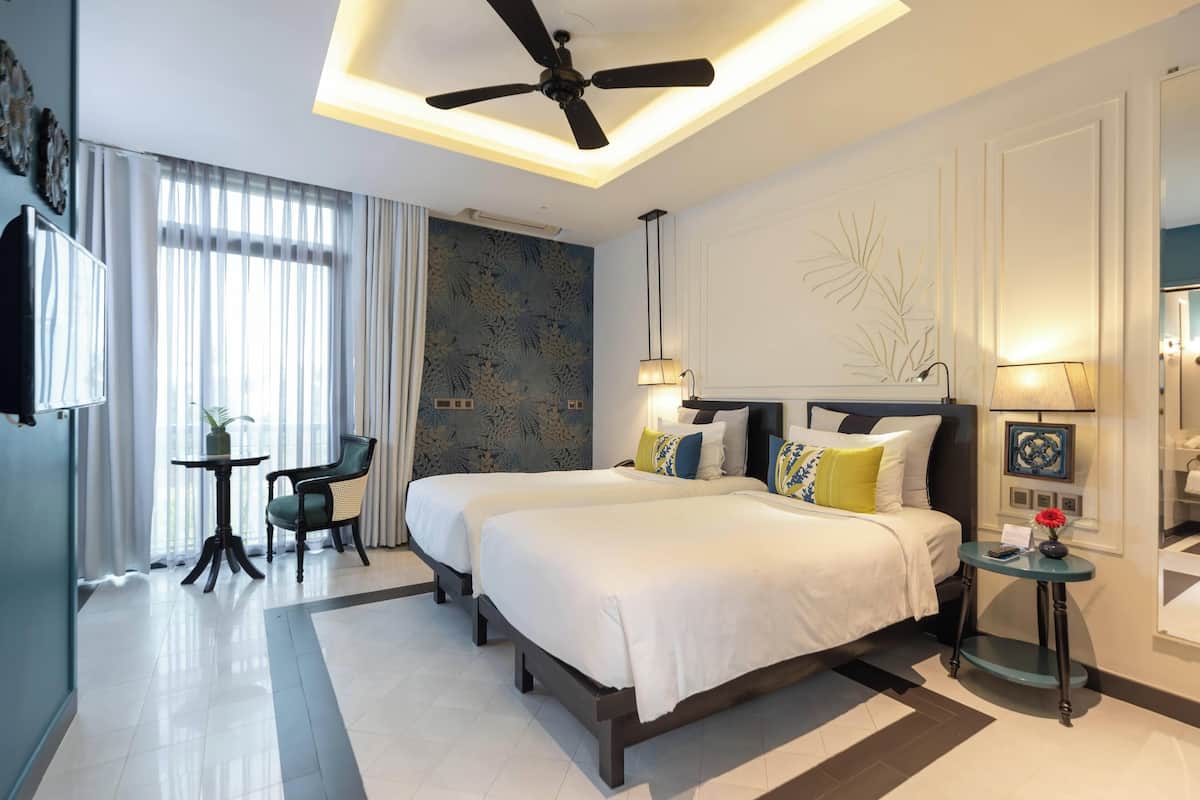 A modern hotel room with two beds, a ceiling fan, a small table with a chair, and a decorative wall piece.