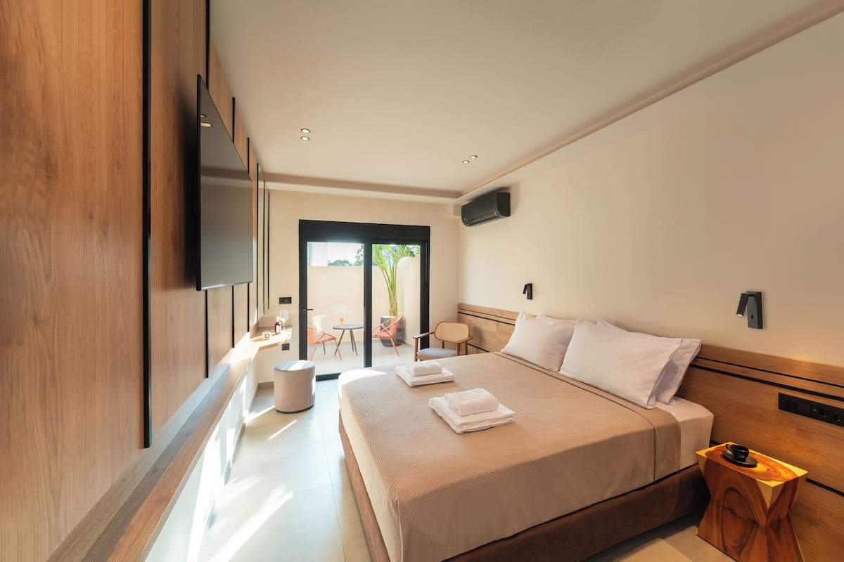 A modern hotel room with a large bed, a flat-screen TV mounted on the wall, a small table with a cup, and a view of the outdoors through a glass door.
