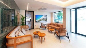 A modern living room with a sofa, armchairs, and coffee tables. There is a large TV mounted on the wall and a window with a view of greenery. - Hippocampus Hotel (Rhodes)