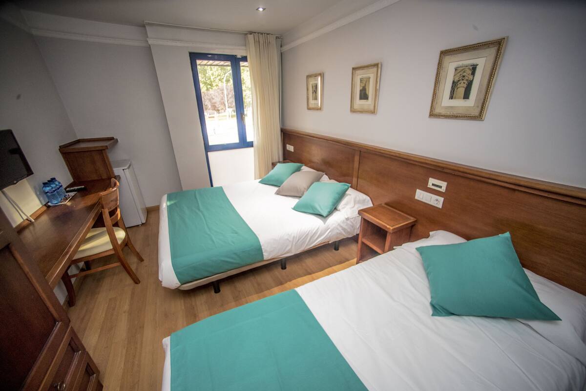A hotel room with two beds, a wooden desk, a chair, and a window with curtains.
