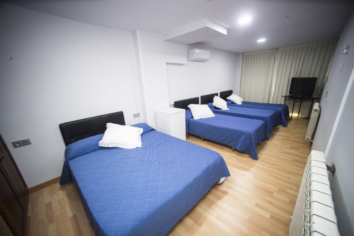 A hotel room with three beds, each with a blue bedspread and white pillows. There is a small white cabinet, a television on a stand, and a radiator on the wall.
