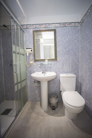 A bathroom with a glass shower enclosure, a white sink, a toilet, and a mirror with a decorative frame. - Hotel Alba (Soria)
