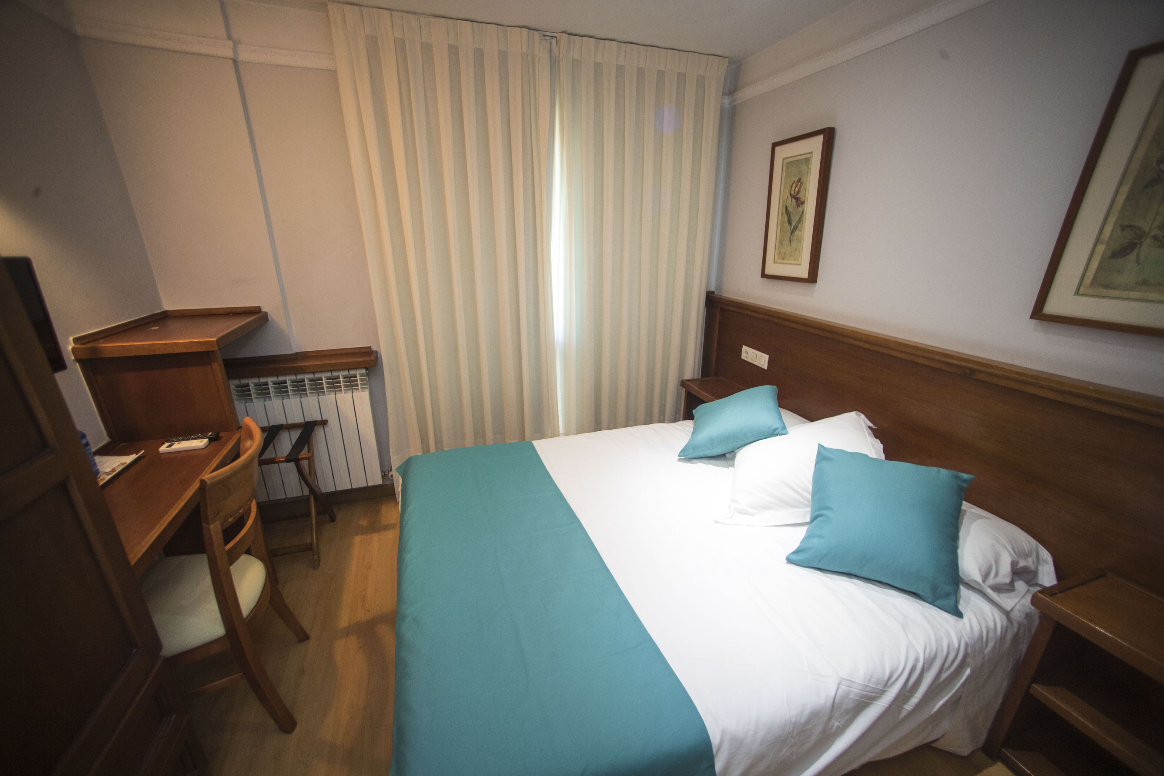 Une chambre d’hôtel avec un lit, un bureau, une chaise, un radiateur et une fenêtre avec des rideaux.