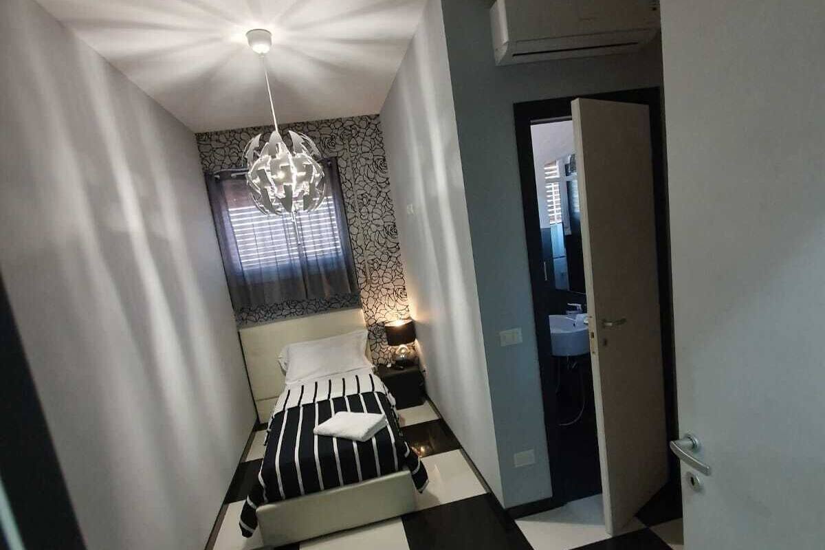 A compact hotel room with a checkered floor, a bed with striped bedding, a small bedside table with a lamp, a window with curtains, and a door leading to a bathroom.