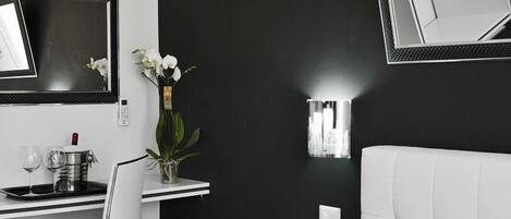 Moderni hotellihuone, jossa on musta tehosteseinä, valkoinen sänky, työpöytä tuolilla, pieni ruukkukasvi ja seinälle asennettu valaisin.