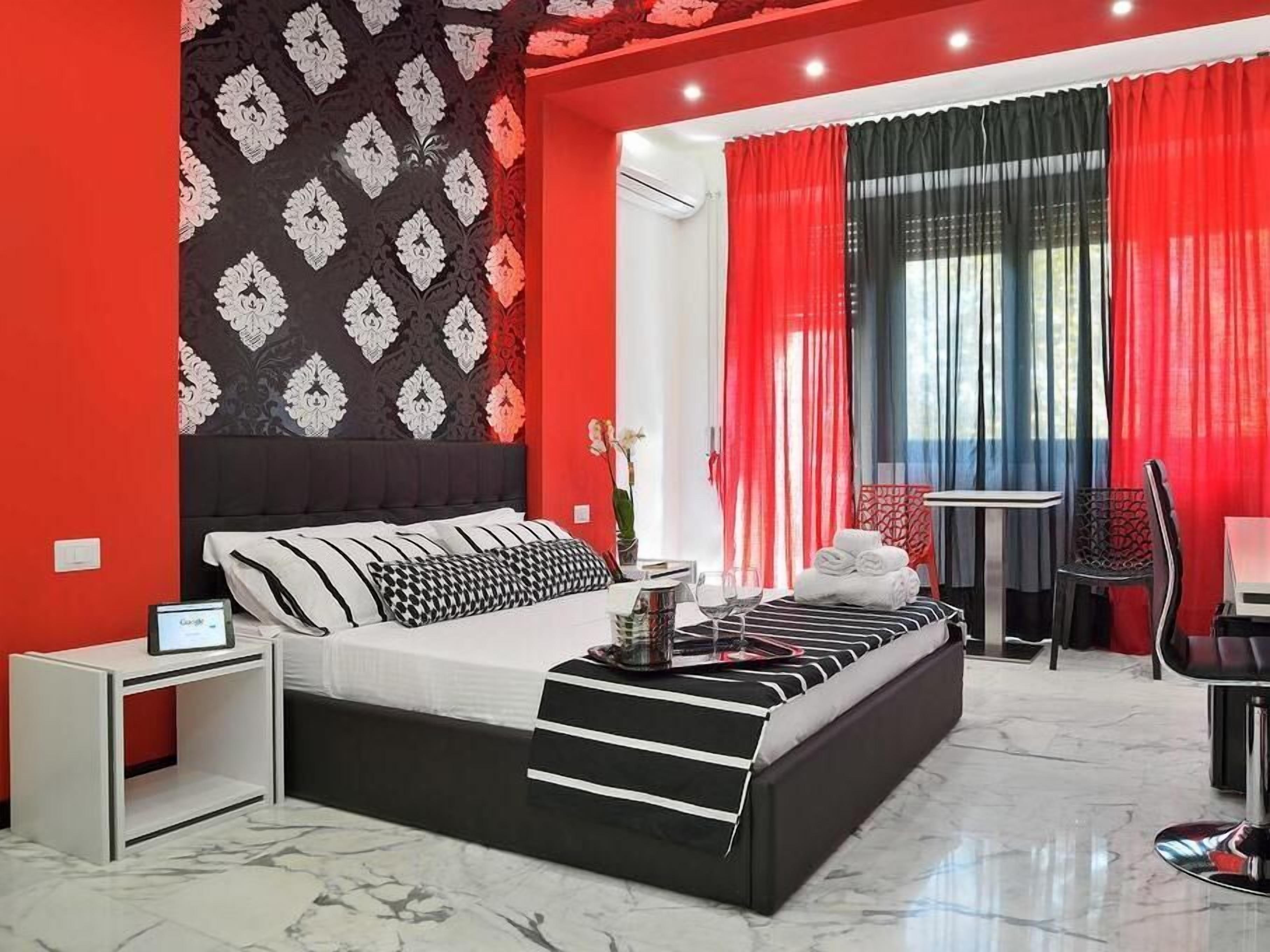 A modern hotel room with a black and white bed, red and black curtains, a small table with a glass, and a chair.