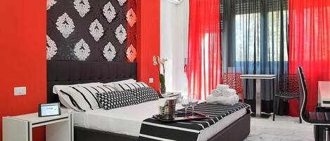 A modern hotel room with a black and white bed, red and black curtains, a small table with a glass, and a chair.