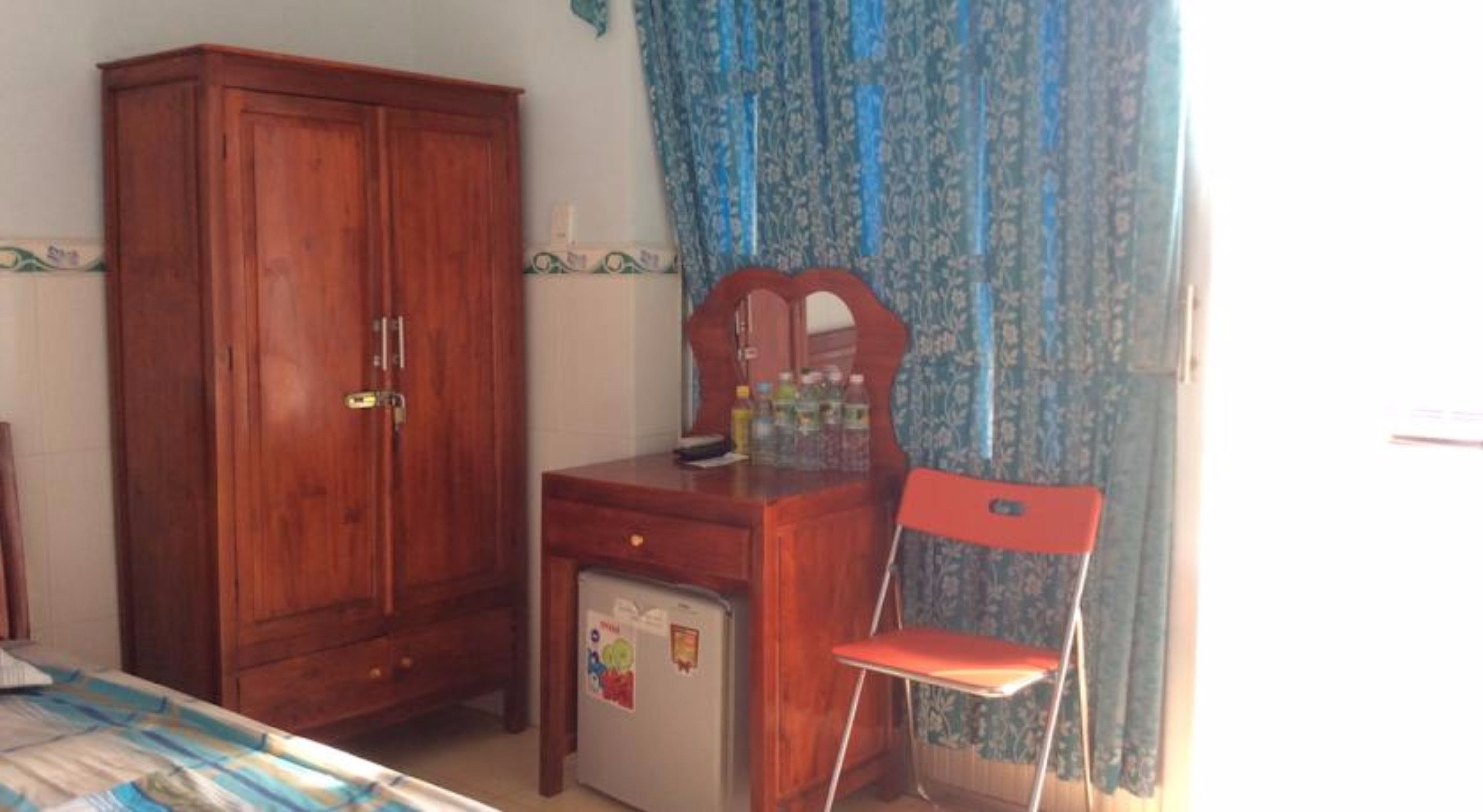 A bedroom with a wooden wardrobe, a small desk with a mirror, a red chair, and a refrigerator.