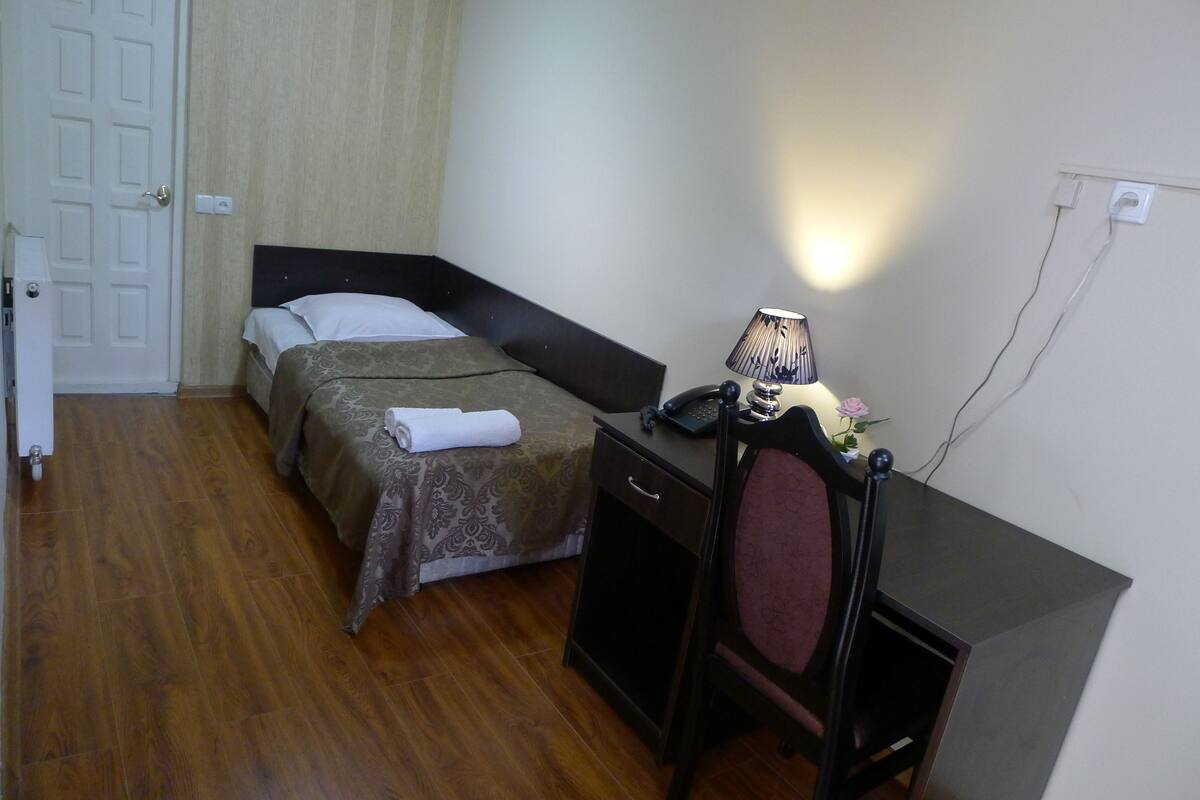 Ett litet hotellrum med en säng, ett skrivbord med en lampa och en stol.