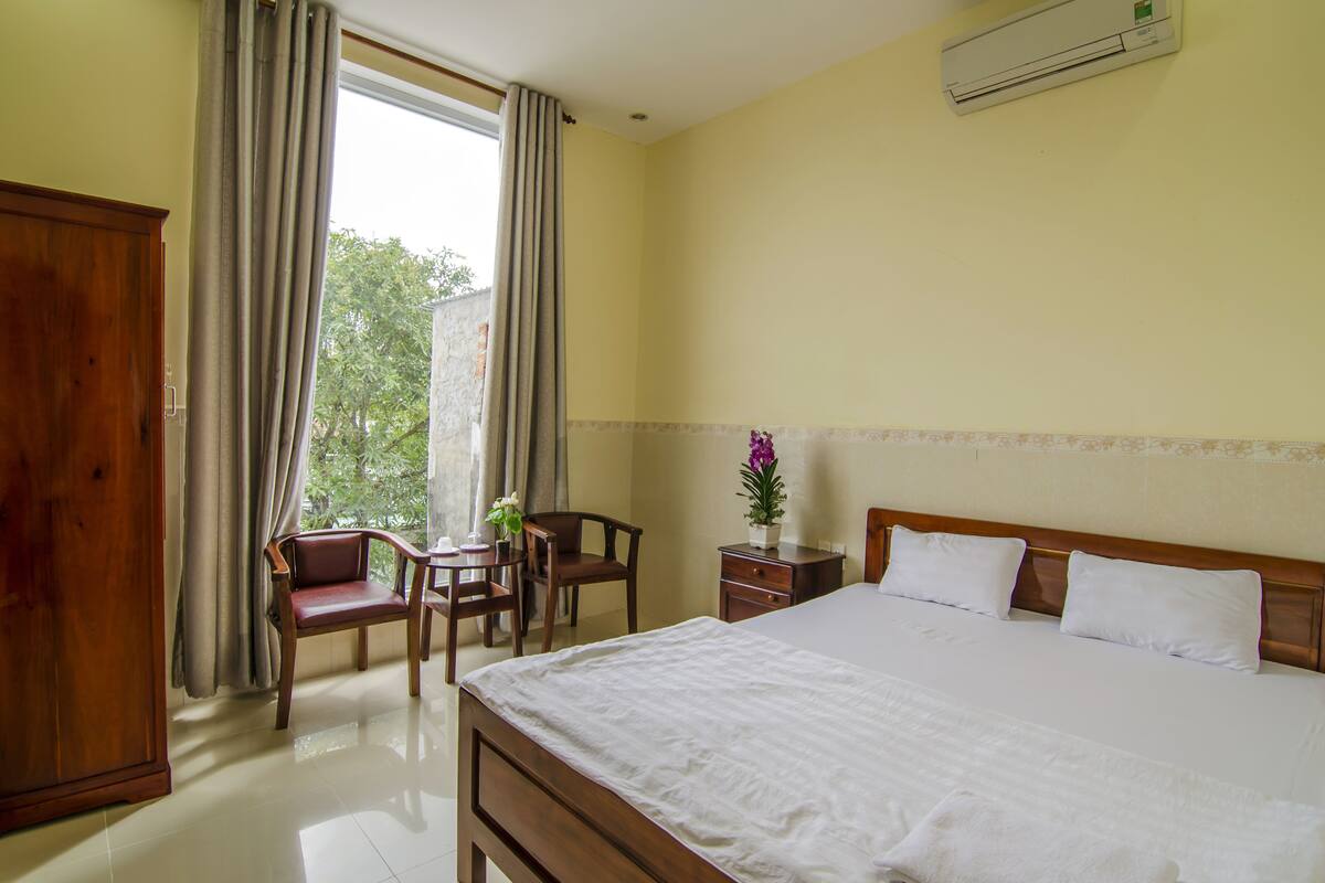 A hotel room with a bed, a wooden wardrobe, a desk with a chair, a small table with a vase of flowers, and a window with curtains.