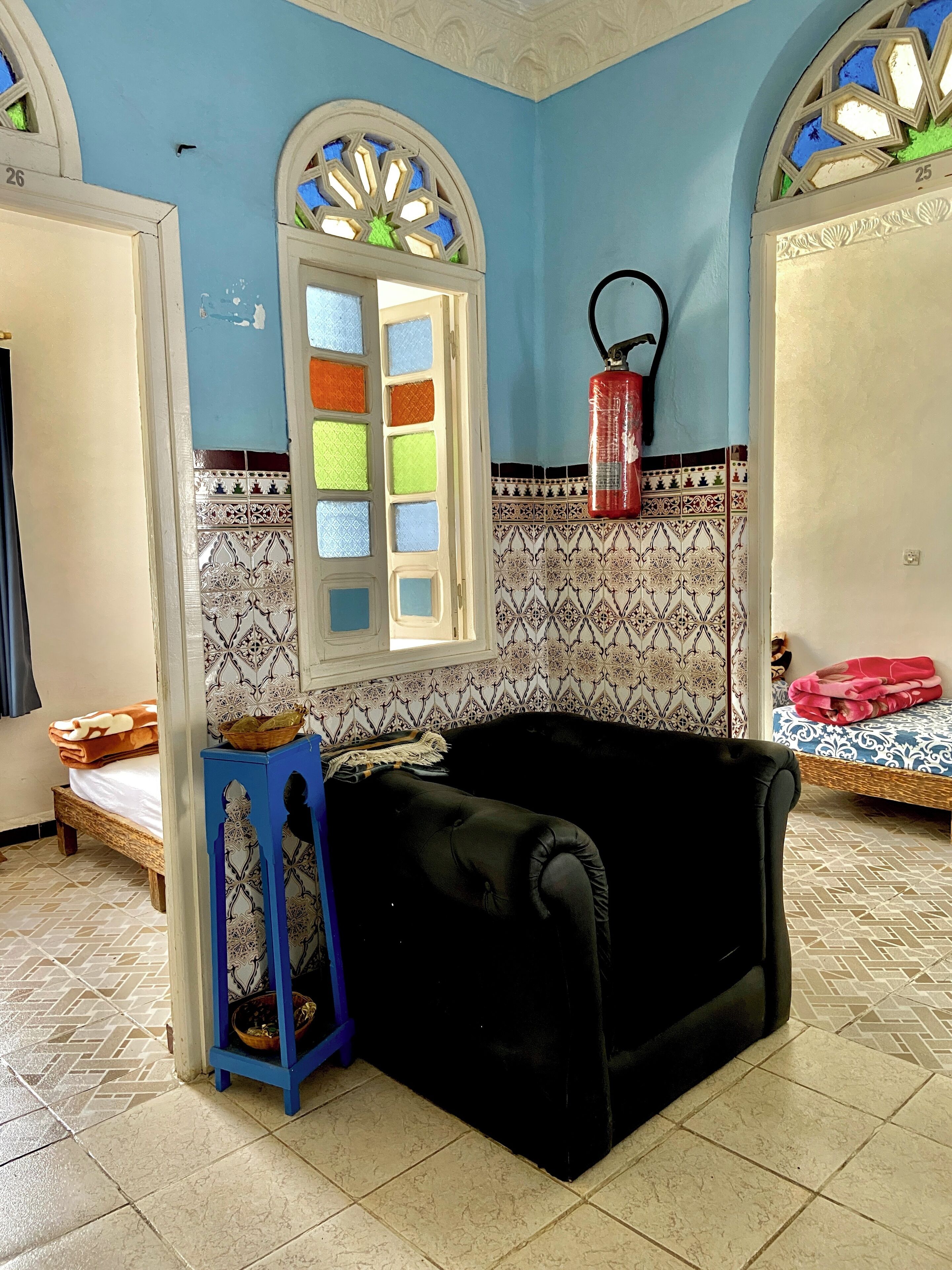 Une pièce avec un canapé noir, un tabouret bleu, un lit recouvert d’une couverture rose et une fenêtre aux vitraux colorés.